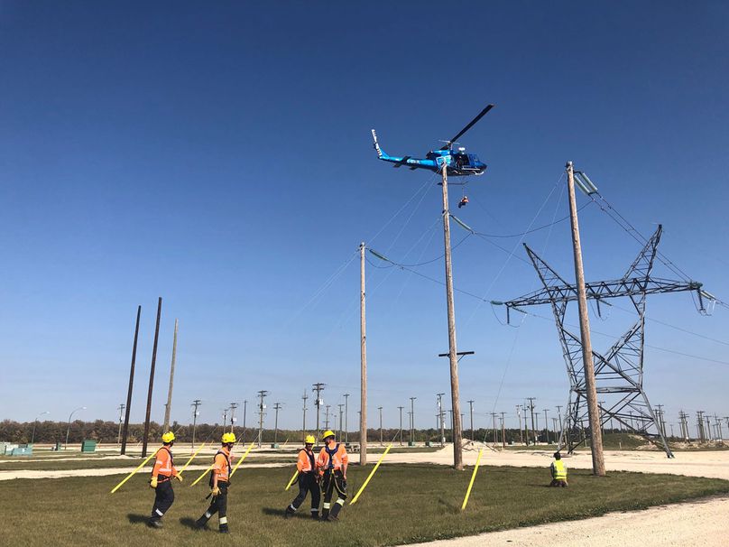 Des monteurs de lignes sous tension debout dans un champ regardent un monteur de lignes suspendu à un câble tiré par un hélicoptère qui survole un terrain d’entraînement.