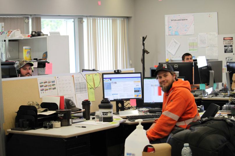 Three men wearing orange high-visibility hoodies sit in different office cubicles in front of computers.