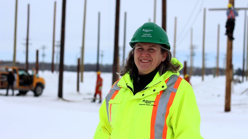 Woman wearing hard hat and safety clothing stands in snowy field as crew climb hydro poles in the background.