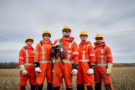 Five power line technicians pose in a field. They are wearing hard hats, orange jumpsuits and safety gloves.