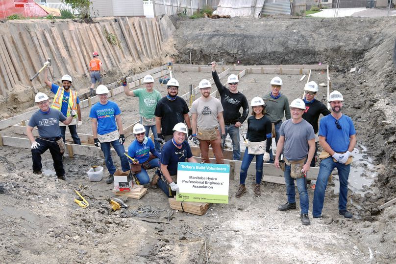 People in hard hats and tool belts stand in an excavated lot beside a framed foundation base.