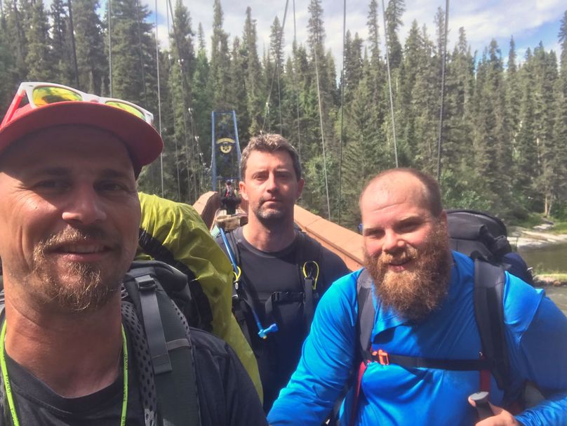 Three men wearing backpacks pose for selfie in boreal forest.