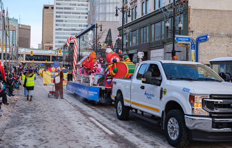 A Manitoba Hydro truck pulls a float with a candy cane, holiday trees, and costumed characters. A reindeer walks beside the truck collecting goods.