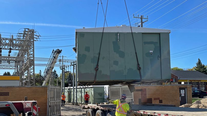 A green building is suspended mid-air by a crane. A worker in high visibility clothing guides it onto a semi- truck trailer.