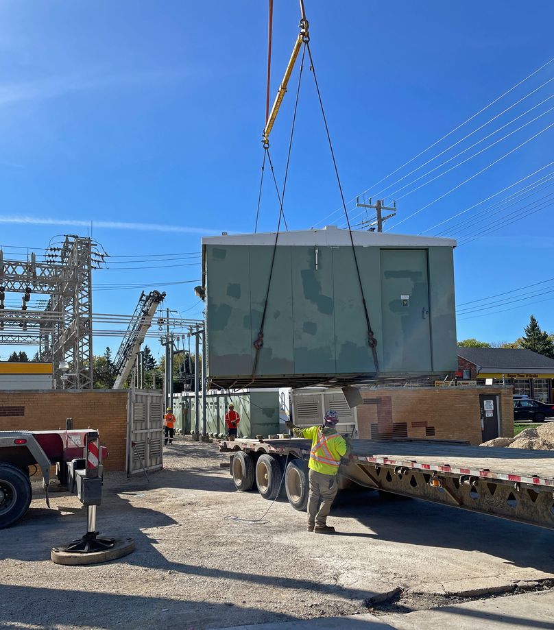 A green building is suspended mid-air by a crane. A worker in high visibility clothing guides it onto a semi- truck trailer.