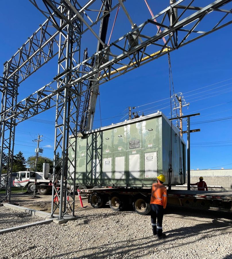 A green building within an electrical substation is hoisted by crane on to a semi-truck trailer