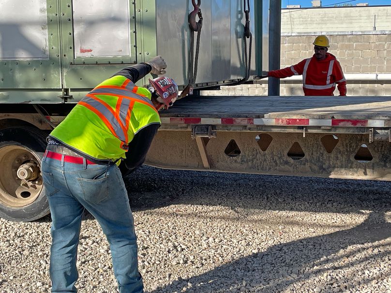 Two workers in high-visibility clothing guide a green building on to a semi-truck trailer.