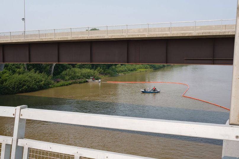 An orange boom trails behind a boat next to a bridge.