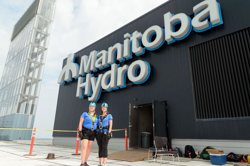 Two women stand on the roof of a building in front of large Manitoba Hydro sign.
