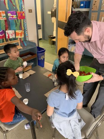 Quatre enfants sont assis autour d’une petite table tandis qu’un homme met des légumes sur du papier absorbant à côté de verres de lait.