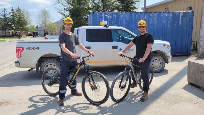 Two men wearing hard hats pose on mountain bikes in front of a Manitoba Hydro truck.