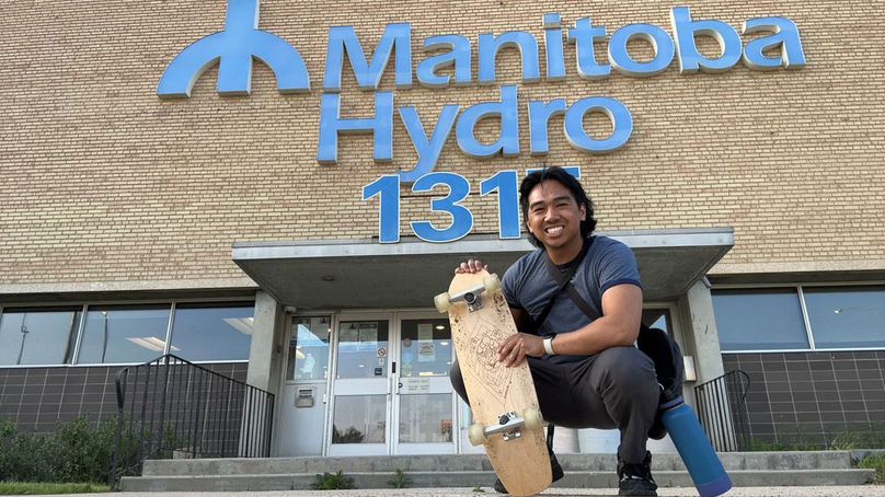 Man crouches with a skateboard in hand in front of a Manitoba Hydro office building.