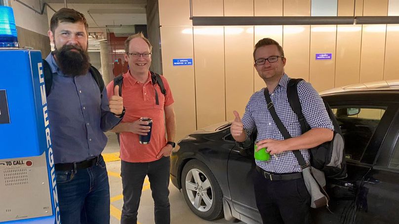 Three people in a parkade show a thumbs up in front of a parking spot labelled “J. Grewal”.
