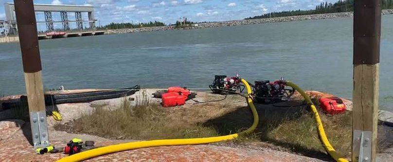 A pair of long yellow hoses connect to gas-powered water pumps sitting on the shore at Jenpeg.