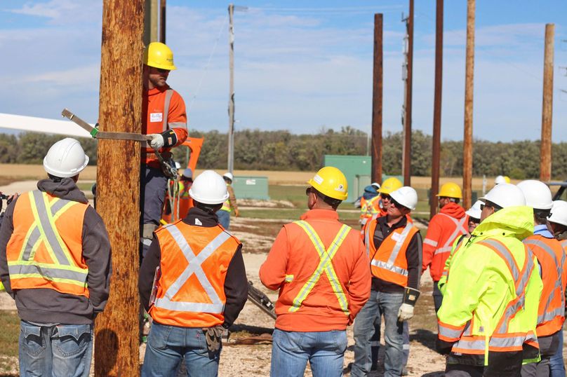 A crowd of people watch a man with climbing gear on a utility pole.