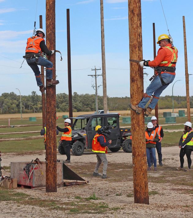 Two men in safety gear climb hydro poles in front of some people.