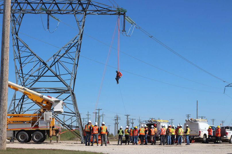 A worker in a harness is suspended from a transmission line high above a large group of people.