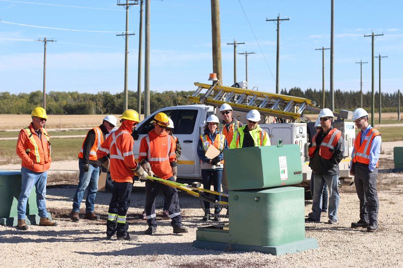 Two male workers use specialized poles to open breakers in a transformer while several people watch.