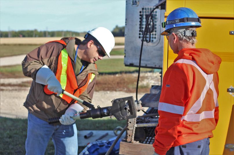 A male worker uses a heavy duty tool to squeeze a pipe while another man watches.