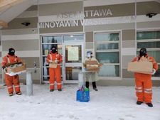Trois employés portant des masques non médicaux tiennent des boîtes remplies de sacs-repas à l’extérieur du centre de santé de Shamattawa (Kisemattawa Minoyawin Waskihekan).