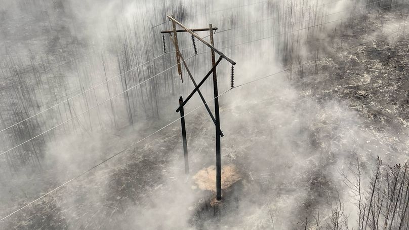 A large transmission line structure made of multiple wooden poles, charred and broken and hanging off power lines amidst a scorched and smoky landscape.
