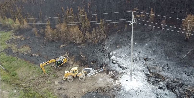 Une vue en plongée d’un camion arrêté près d’un poteau de ligne de transmission brûlé par un incendie échappé. La forêt derrière le poteau est brûlée à certains endroits, mais de la végétation subsiste.