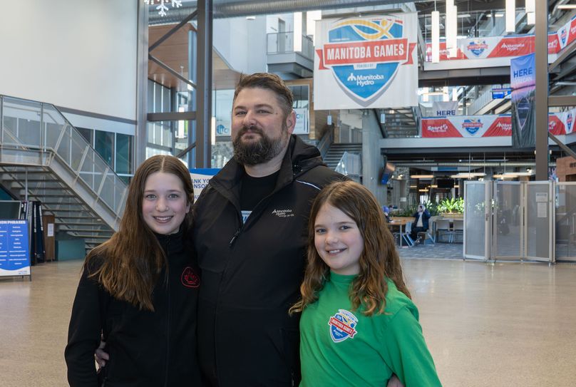 A man and his two daughters pose in a large lobby with many Manitoba Games signs.