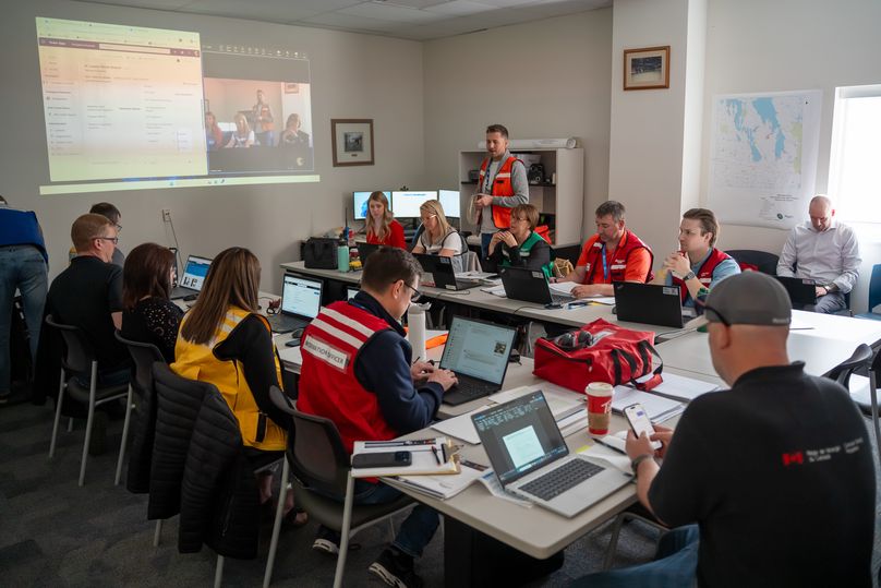 A group of people with laptops sit in a circle facing a projector screen.