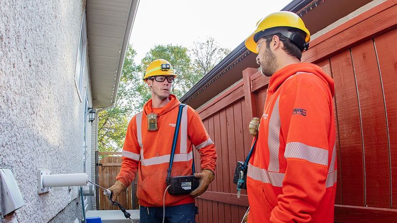 Two men in high visibility clothing chat while using a handheld monitoring device on an exhaust pipe outside a residence.
