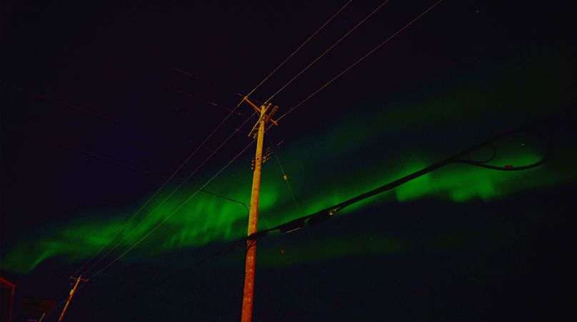 Les lueurs boréales (Aurora Borealis) dansent dans le ciel nocturne, mettant en valeur les tons vifs de vert. Un poteau électrique est en toile de fond.
