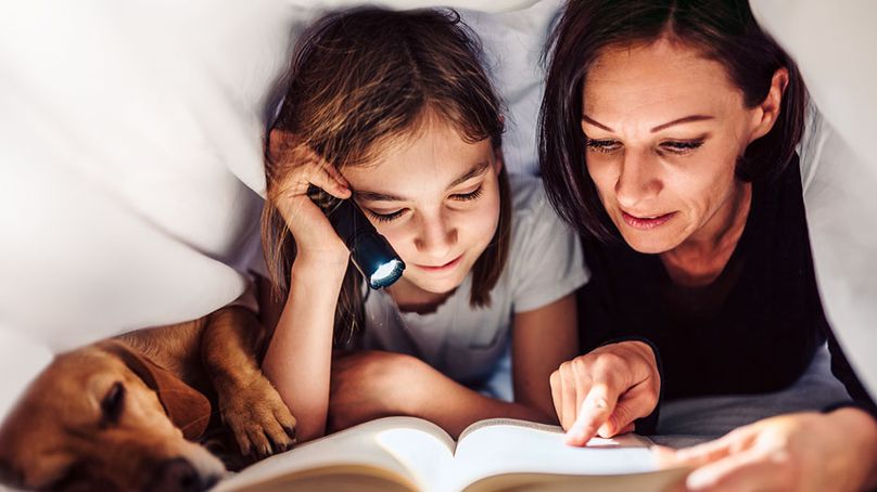 A mother and daughter read under the covers by flashlight together. Their dog sleeps beside them.