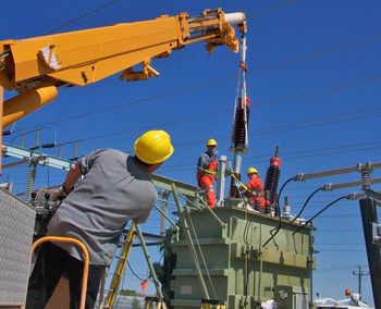 A man uses a crane to lift electrical equipment up to two people working on a transformer.