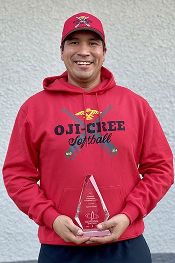 A smiling man, wearing a red Oji-Cree Softball sweater and baseball cap, holding a glass award.