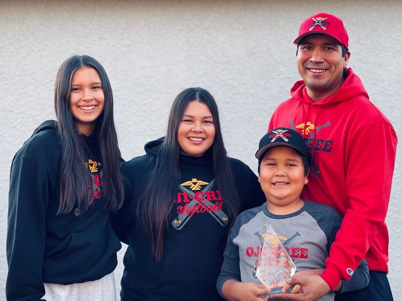 Une fille, une femme, un homme et un garçon qui sourient, tous vêtus de chandails d’Oji-Cree Softball, et qui présentent un prix en verre.