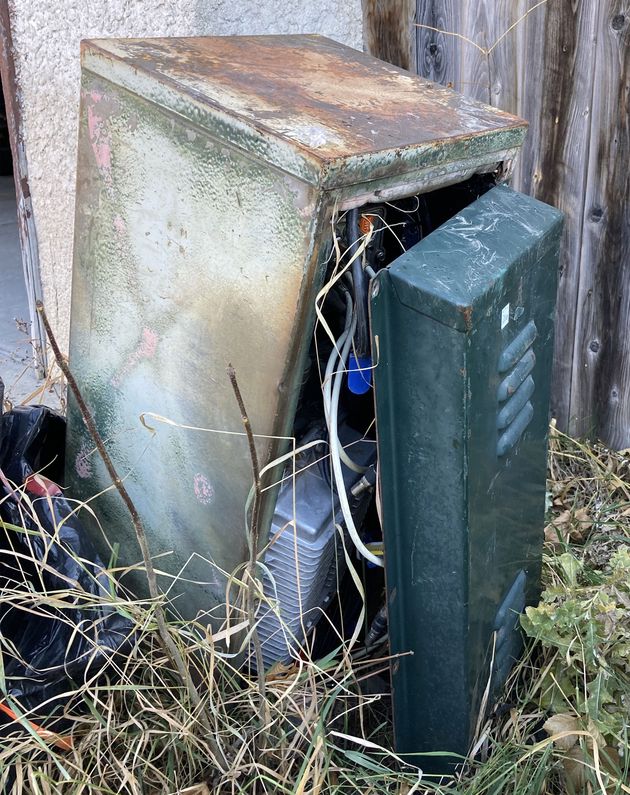 Photo d’une boîte métallique verte, à la hauteur du genou, se trouvant dans de hautes herbes à côté d’une clôture. La boîte, rouillée, penche d’un côté; le panneau de droite présente des évents au haut et au bas, et est séparé du reste de la boîte. Le diamètre des fils qui sortent de la boîte ne dépasse pas celui d’un crayon.