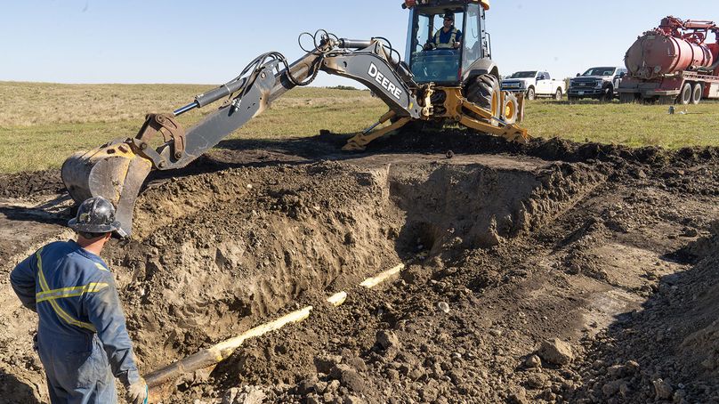 A front-end loader digs up soil to expose an underground gas pipe. Another worker stands beside the pit with a shovel.