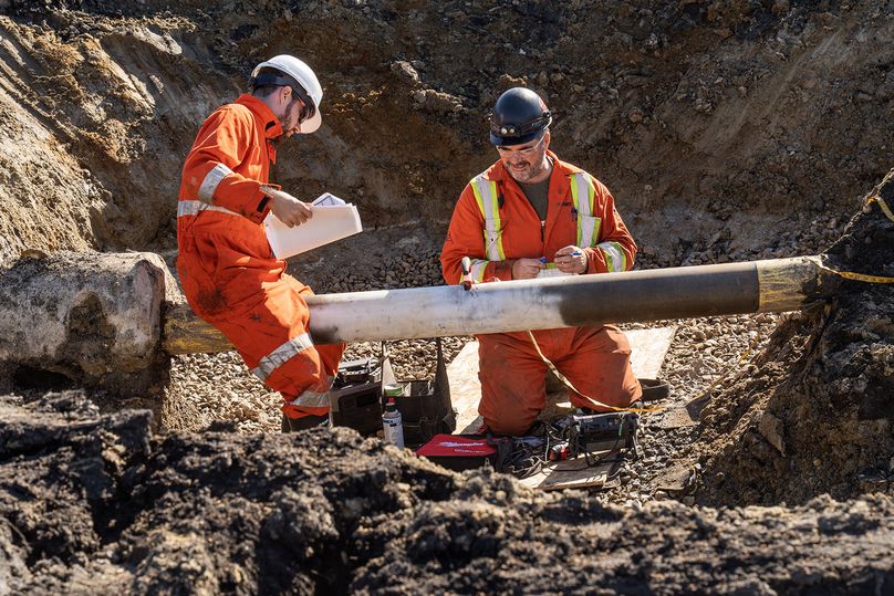 Two men sit in a trench, examining an exposed underground gas pipe.