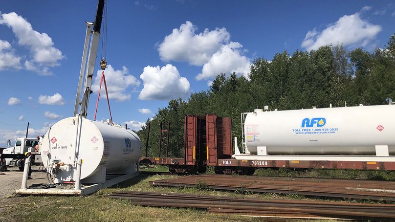 A crane moving a large metal fuel tank off a train car and onto the ground nearby.