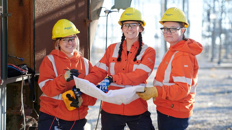 Three women in safety gear stand outdoors and consult over some planning drawings at a Manitoba Hydro facility.