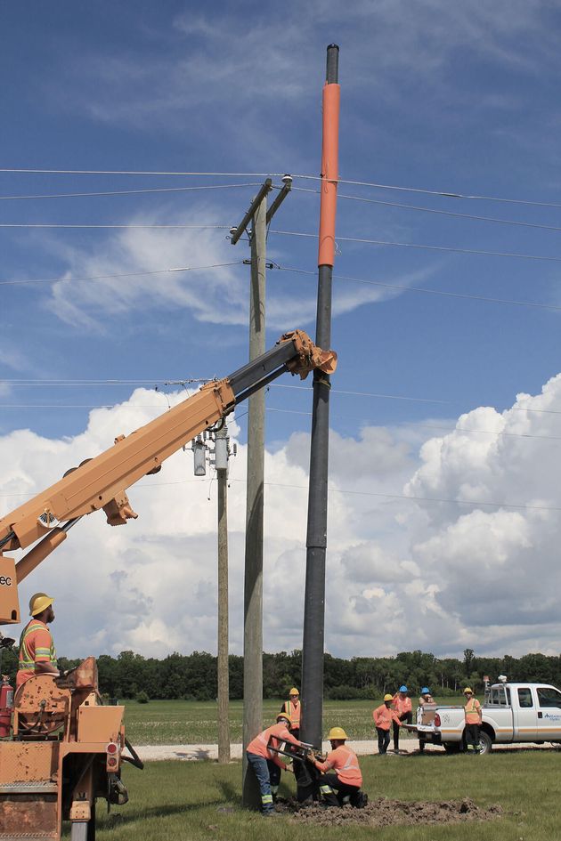 A composite/fibreglass pole that has been assembled is being stood up by a crane arm and employees work on the base