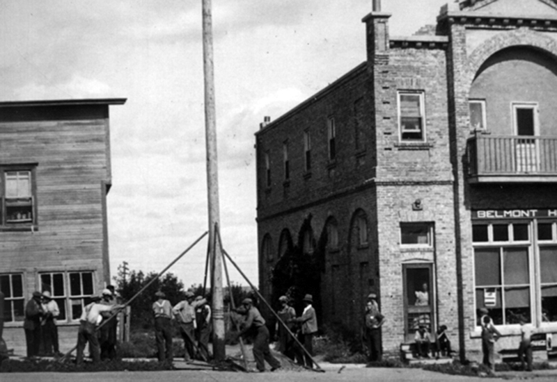 Workers erecting a wooden hydro pole beside the Belmont Hotel while townspeople look on.