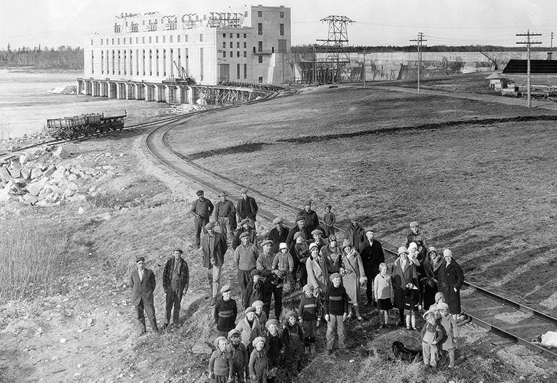 Employees, their spouses, and children stand beside the railway line outside of Great Falls generating station. The generating station is in the background.