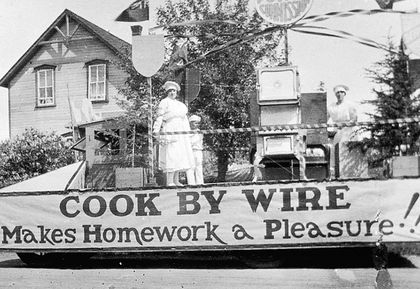 Un char allégorique à Carman avec trois personnes en tenue de chef (chapeaux et tabliers) exposant fièrement une cuisinière électrique. Sur la banderole on lit : Cook by wire makes homework a pleasure!