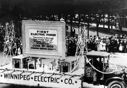 Un char allégorique de la Winnipeg Electric Co. dans un défilé, passant devant des foules. Le char, formé de bâtiments et de pylônes de transmission, marque la transmission de Pinawa à Winnipeg.