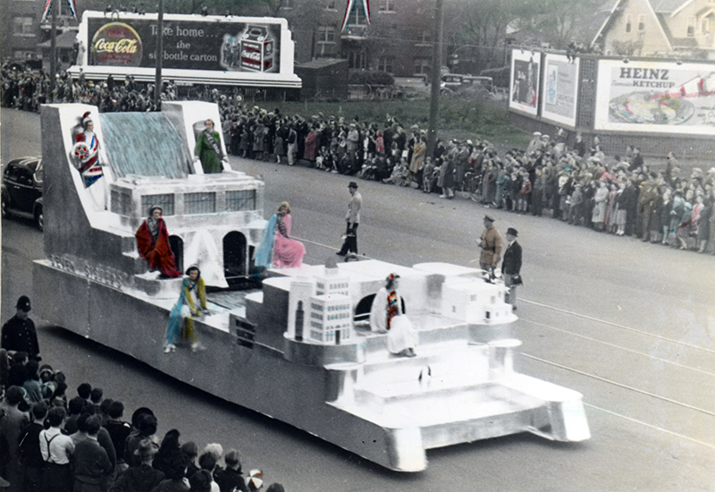 People in fancy costumes sitting on a parade float representing a hydroelectric dam and a city that receives its electricity. People line both sides of the street.